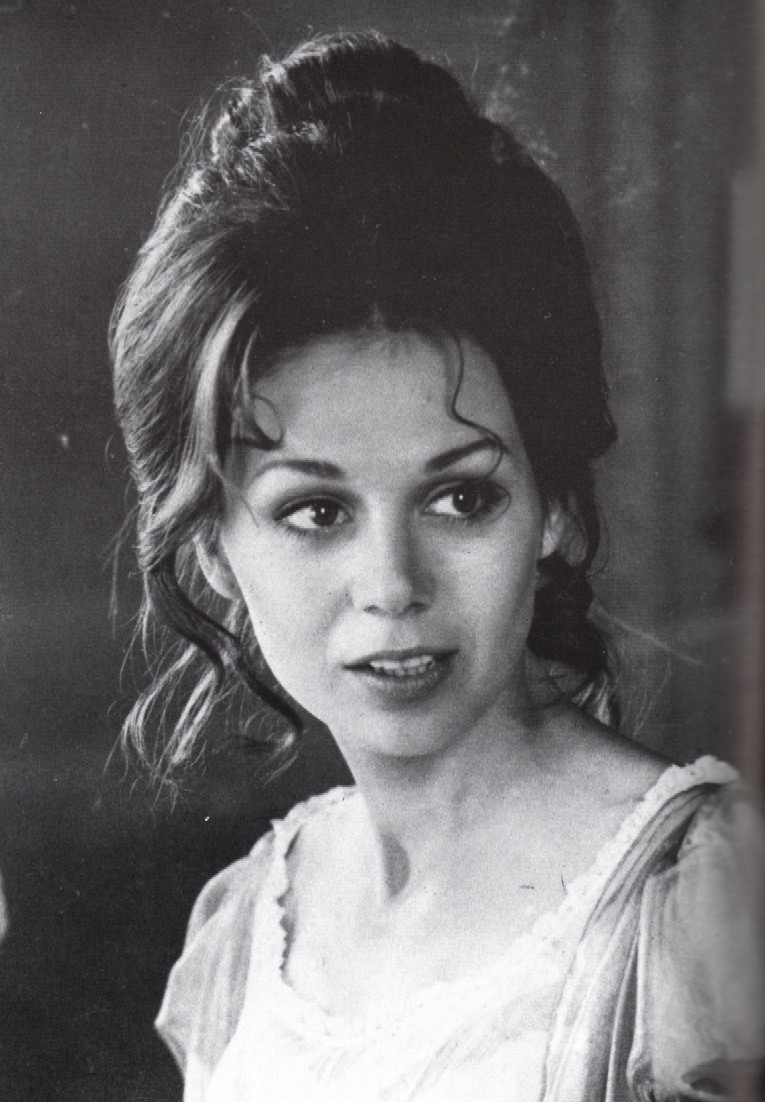 A black and white portrait image of Maggie. She has pale skin, a white dress, and brown hair in an updo.